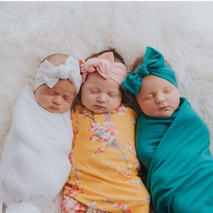 Three newborns wrapped in soft swaddle blankets with matching floral headbands on plush rug