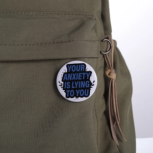 Close-up of a tinplate badge with 'YOUR ANXIETY IS LYING TO YOU' on a green canvas bag