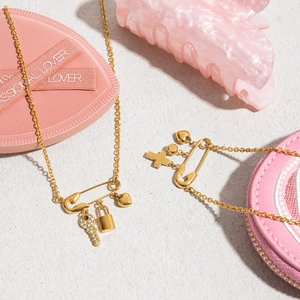 Detail of gold paperclip necklaces with cross, key, and lock charms on a soft pink surface