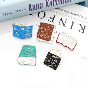 Five assorted book-shaped enamel pins with gold borders on white ceramic plate
