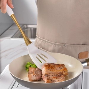 White silicone slotted turner with gold handle flipping meat in a frying pan
