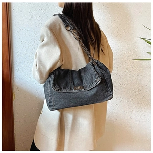 Model wearing dark denim tote bag with metal chain strap and flap design against a wall