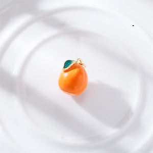 Macro shot of an orange enamel fruit pendant with green leaf and gold loop on ceramic