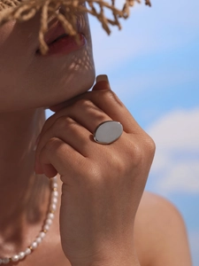 Close-up of a silver oval open ring worn by a model against a blue sky beach background