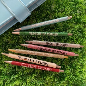 Flat lay of seven glittery Christmas weekday pens on green grass with a blue folder
