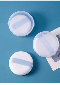Three round white velvet powder puffs with blue ribbon handles in a clear storage box