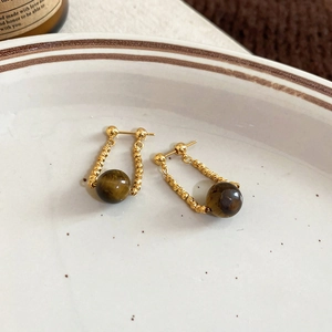 Dettaglio di orecchini a U dorati intrecciati con pietre occhio di tigre su fondo ceramica