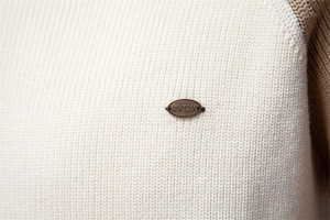 Detailed macro view of cream rib-knit texture with brown seam and metal brand plaque