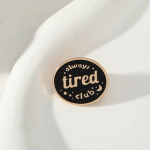 Black oval enamel pin featuring always tired club text with celestial stars and moon gold trim