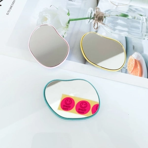 Three pebble-shaped handheld makeup mirrors in pink, yellow, and cyan frames on a desk