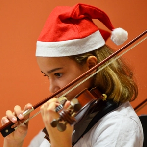 Close-up of a child wearing a red Santa hat while playing a brown violin on orange background