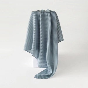 Detailed view of greyish-blue pleated scarf draped on white block in minimalist studio
