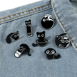 Set of seven cartoon black cat enamel pins displayed on blue denim fabric background