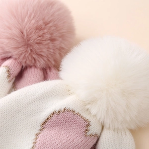 Macro detail of infant knit hat featuring pink yarn, gold lurex accents and fluffy pom-poms