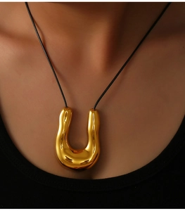 Close-up of a large golden U-shape geometric pendant on a black braided leather cord necklace
