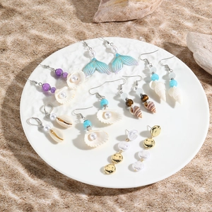 Flat lay of ocean-themed shell and mermaid tail earrings with pearls on sand texture