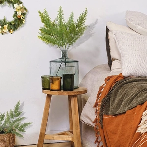 Artificial pine branches in a green glass vase on a wooden stool with Christmas wreath