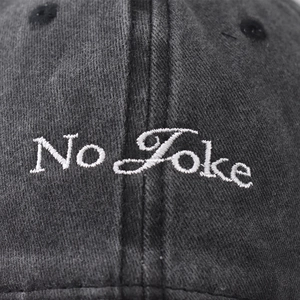 Close-up of white No Joke embroidery on washed black cotton denim baseball cap