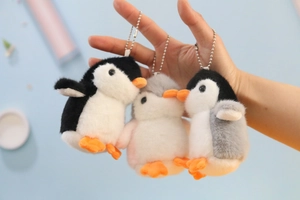 Three cute plush penguin keychains with metal bead chains held by hand against blue backdrop