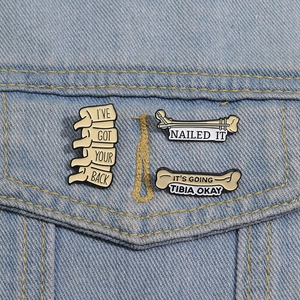 Three enamel pins with medical puns on denim: I've Got Your Back, Nailed It, Tibia Okay