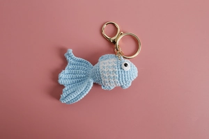 Close-up of blue handmade crochet goldfish keychain with golden ring on pink background