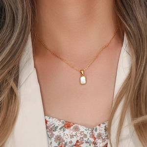 Detail of a gold necklace with a square white shell pendant worn by a model in a beige blazer