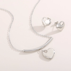 Detail view of silver heart pendant necklace and stud earrings set on soft pink backdrop