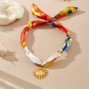 Vue détaillée du collier foulard en soie coloré avec pendentif soleil doré sur le sable