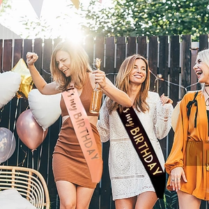 Pink and black satin birthday sashes with gold lettering in festive outdoor party setting