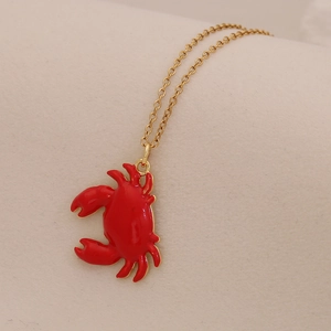 Detail of a golden necklace featuring a vibrant red enamel crab pendant on beige fabric