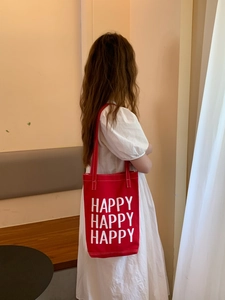 Detail of red canvas tote bag with white HAPPY text print worn by model in white dress