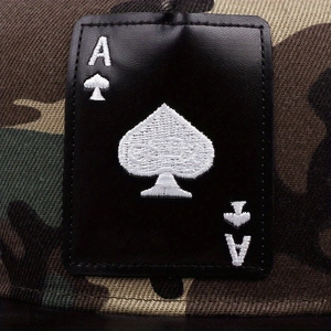 Gros plan d'un écusson en cuir noir brodé d'un As de Pique sur une casquette camouflage