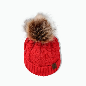 Detail of red cable knit beanie featuring a brown faux fur pom and leather label accent
