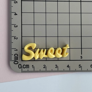 Détail d'un charme Sweet en résine jaune sur tapis de découpe avec règle de mesure