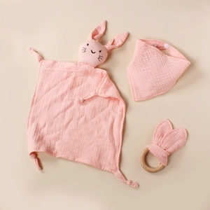 Detail of pink cotton muslin baby bib, wooden rabbit ear rattle and plush comforter set