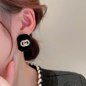 Close-up of black velvet square earrings with gold rhinestone logo worn by a model