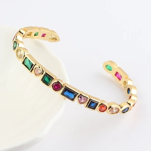 Gold open bangle with colorful geometric zircons displayed on a minimalist white ceramic bowl
