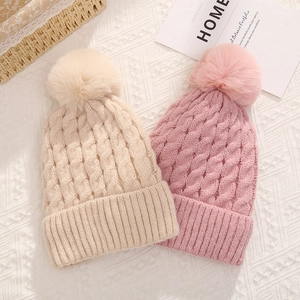 Off-white and pink cable knit beanies with large faux fur pom poms on textured fabric
