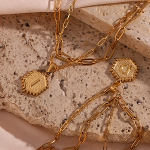 Macro detail of golden paperclip chains and hexagonal initial pendants on stone surface