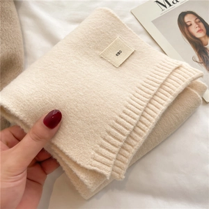 Close-up of a soft beige knitted scarf featuring a black emo logo tag held by hand