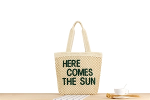 Détail du sac cabas en paille tissée avec broderie verte Here Comes The Sun sur table