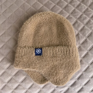 Dettaglio cappello in maglia cammello con paraorecchie, ecopelliccia e fiocco di neve