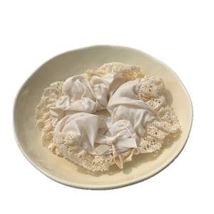 Elegant cream silk scrunchie with lace trim, satin bow, and shell charm in a ceramic bowl