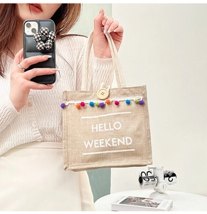 Detail of jute tote bag showing Hello Weekend text, wood button, and colorful pompoms
