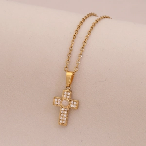 Dettaglio di un ciondolo a croce dorata con zirconi micro-pave su uno sfondo beige testurizzato