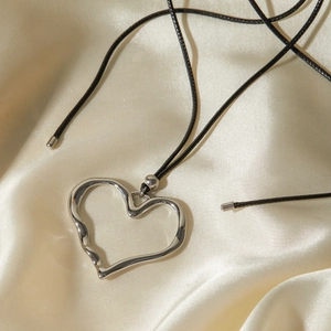 Silver hollow heart pendant with black wax cord on beige silk fabric background
