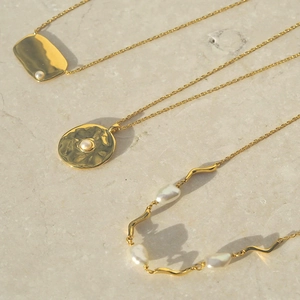 Three gold baroque pearl necklaces with hammered metal pendants on a marble surface
