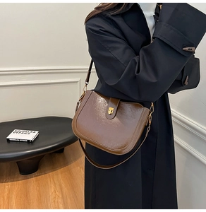 Model wearing dark brown glossy leather saddle bag with gold buckle over black trench coat