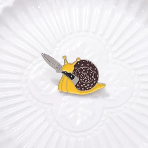 Cartoon enamel pin featuring a cute yellow snail holding a silver dagger with a brown shell