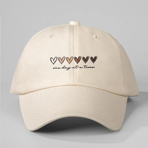 Detail of cream baseball cap featuring gradient hearts and 'one day at a time' text